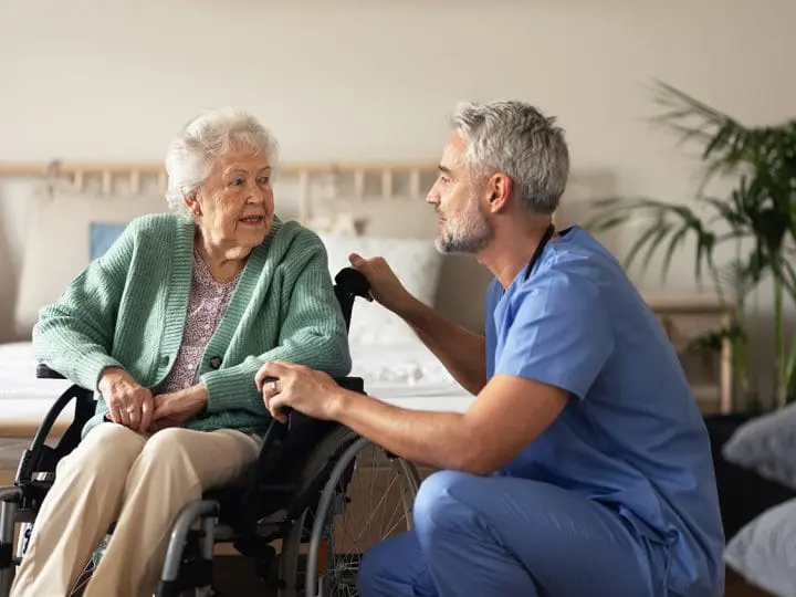 A caregiver kneels down to talk with a woman with late-stage dementia.
