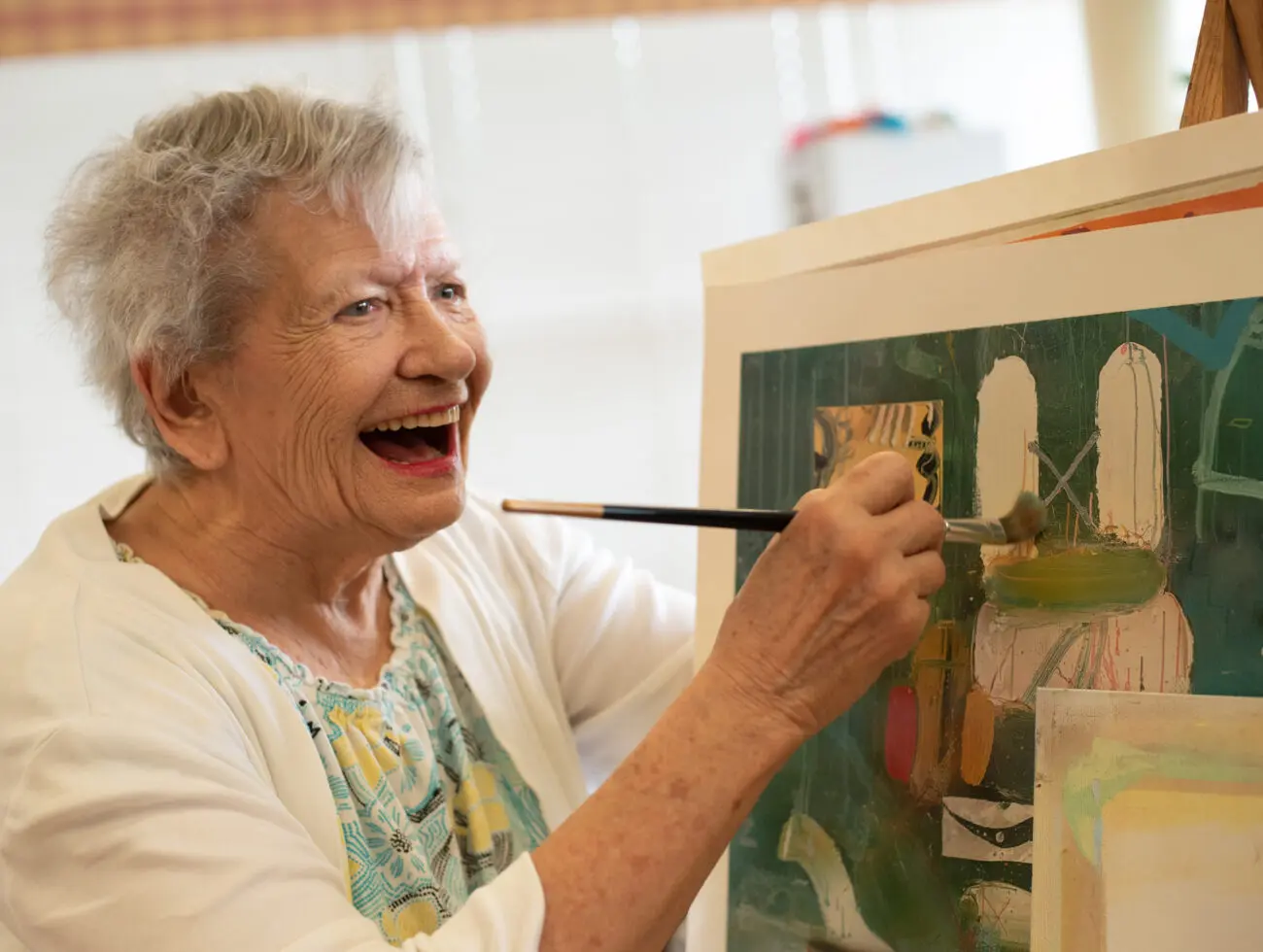A senior woman painting on a canvas while smiling, showcasing creative art therapy.