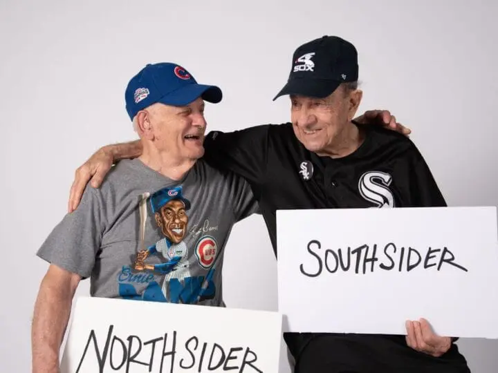 Two elderly men pictured together and holding signs that read, "south sider" and, "north sider", showcasing acceptance.