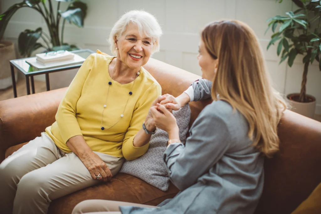 senior woman holding hands with woman next to her on couch in Central Baptist Village, as they connect over fun questions to ask someone with dementia.
